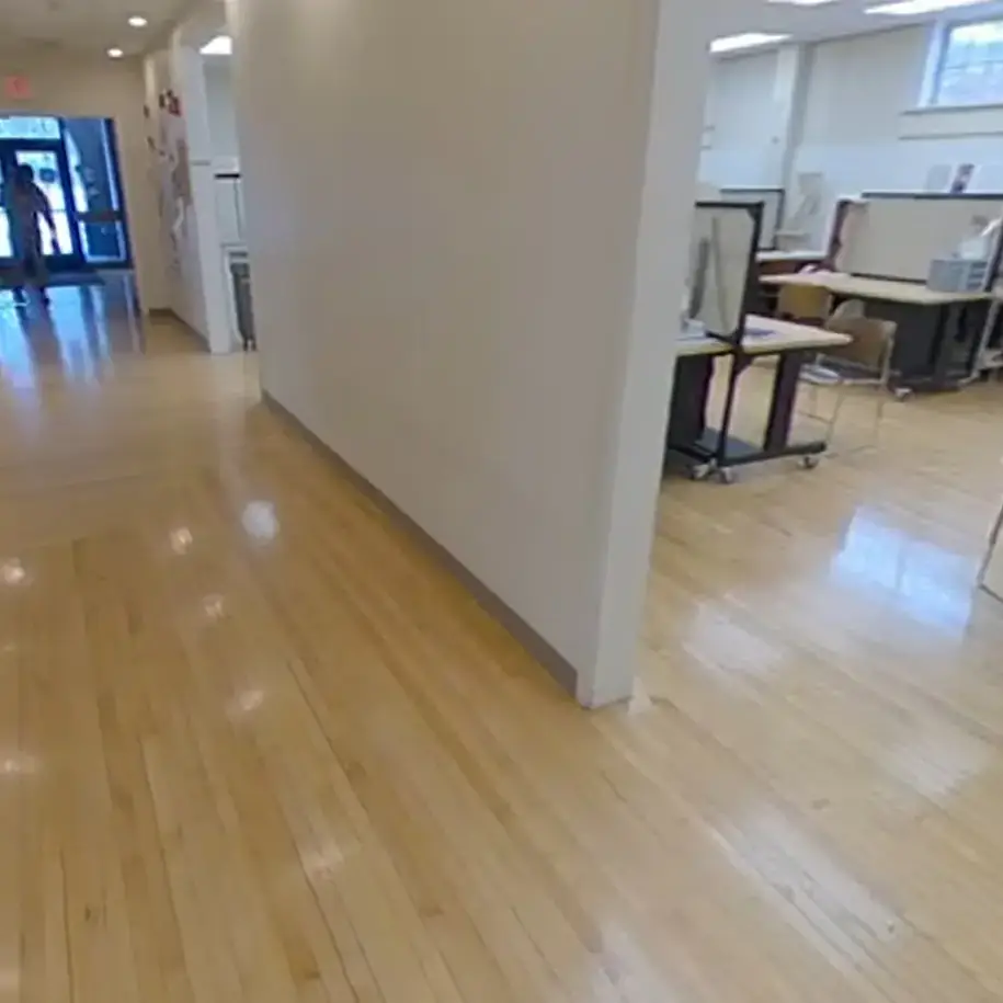 Windowed room with desks and dividers, and hallway separated by a wall and archway, with shiny wooden floors.