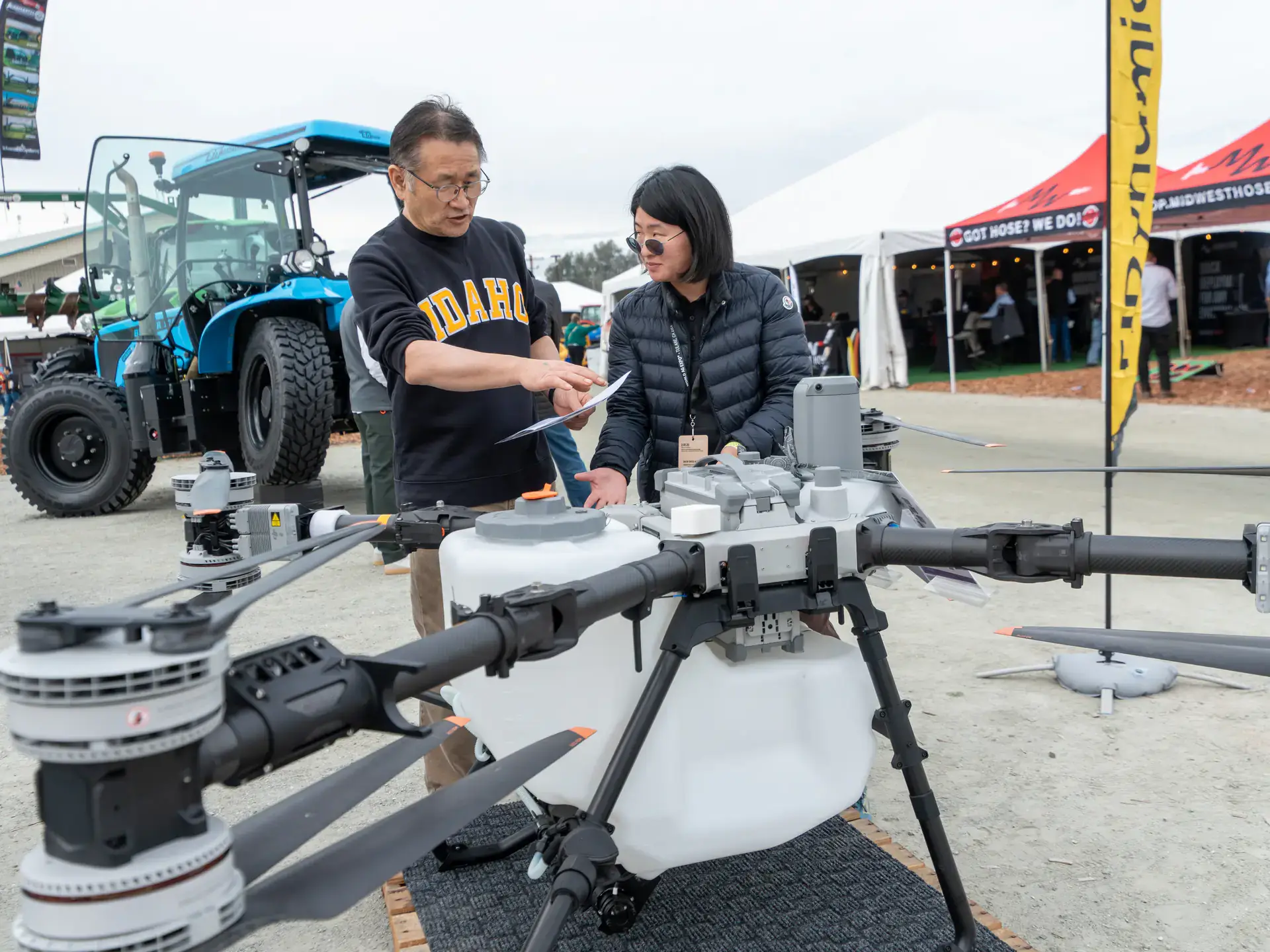 A man and woman standing in front of a farm tractor discuss a large drone in front of them. 