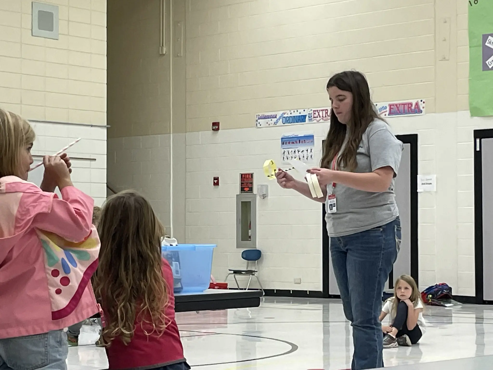  A woman standing inside of a school gymnasium holds a plane made from a straw and paper while children watch attentively.