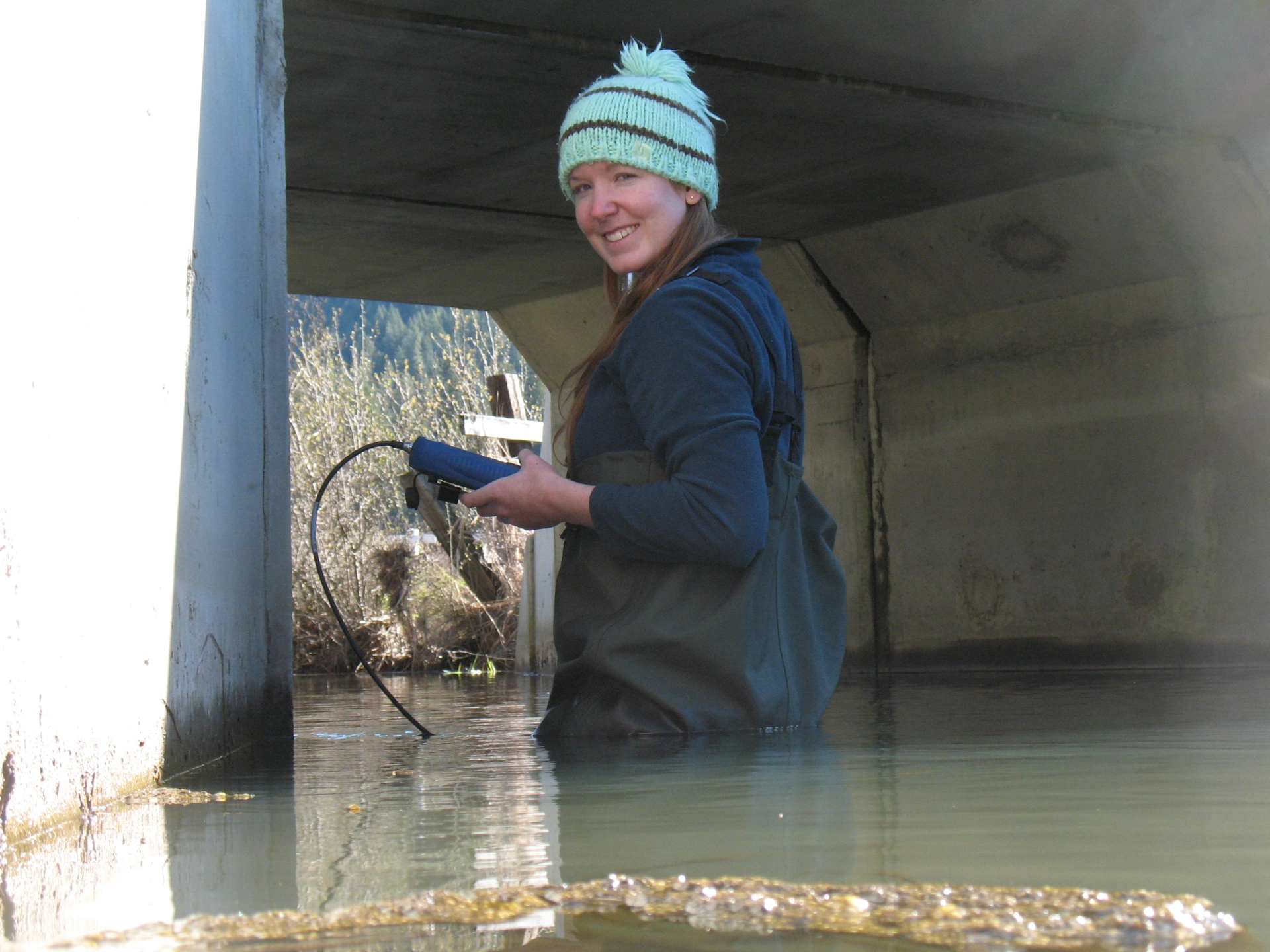 Trea LaCroix wades in a lake wearing waders and takes measurements under a bridge. 