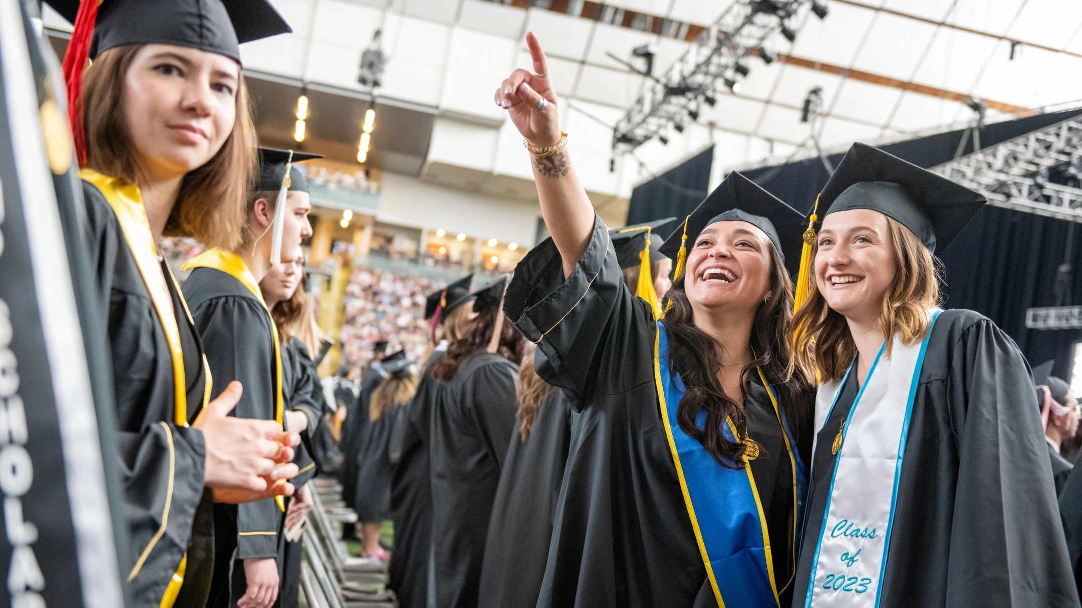 Two graduating students in their cap and gown, with one pointing to the bleachers