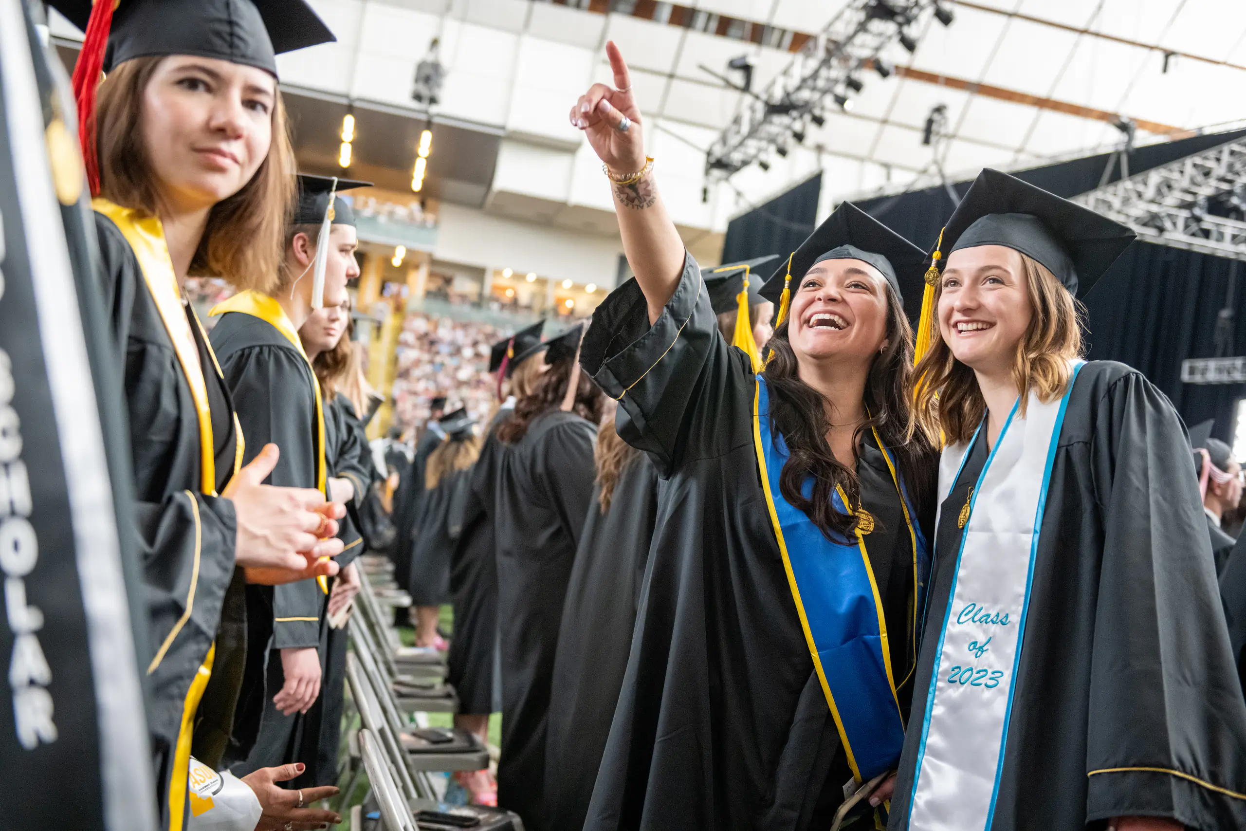 Two graduating students in their cap and gown, with one pointing to the bleachers
