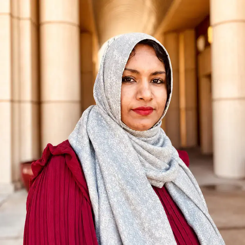 A woman wearing a red dress and a gray headscarf poses in front of a columned architectural structure.