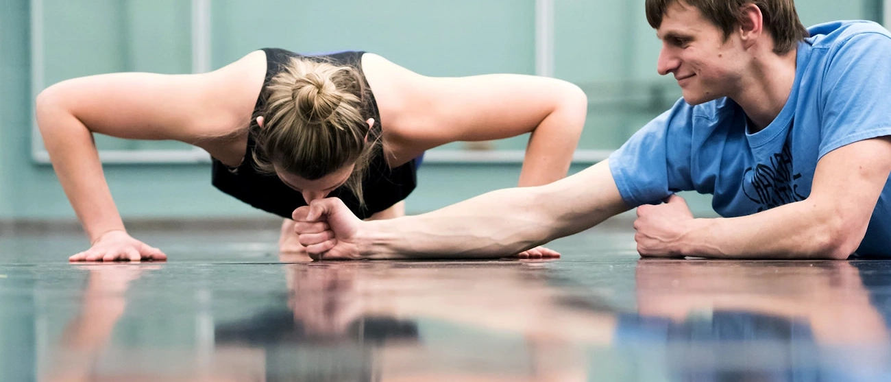 Woman doing pushup with man holding a fist to her forehead on the floor.