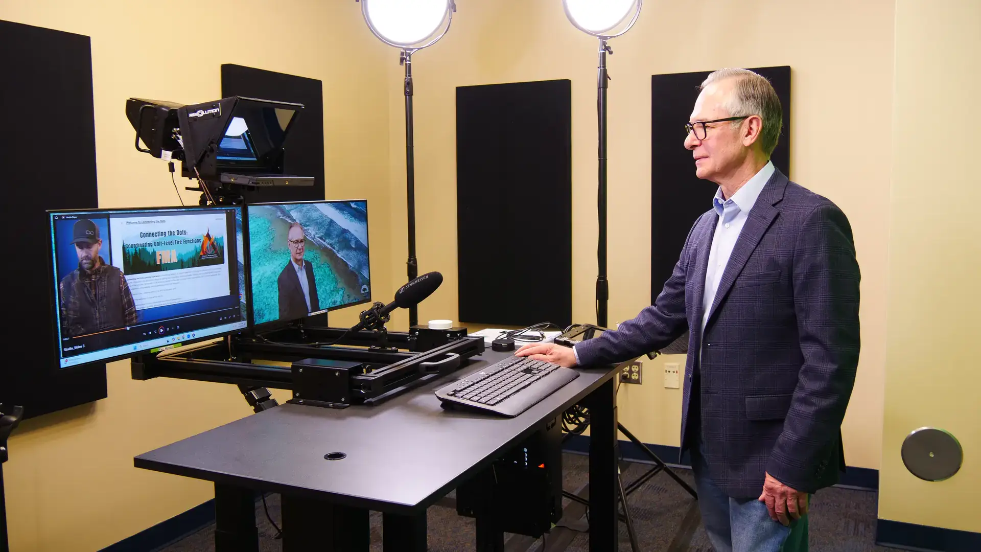 Rick Gividen, wearing a blue sport coat and jeans, stands at a multimedia desk with screens, microphone and teleprompter.