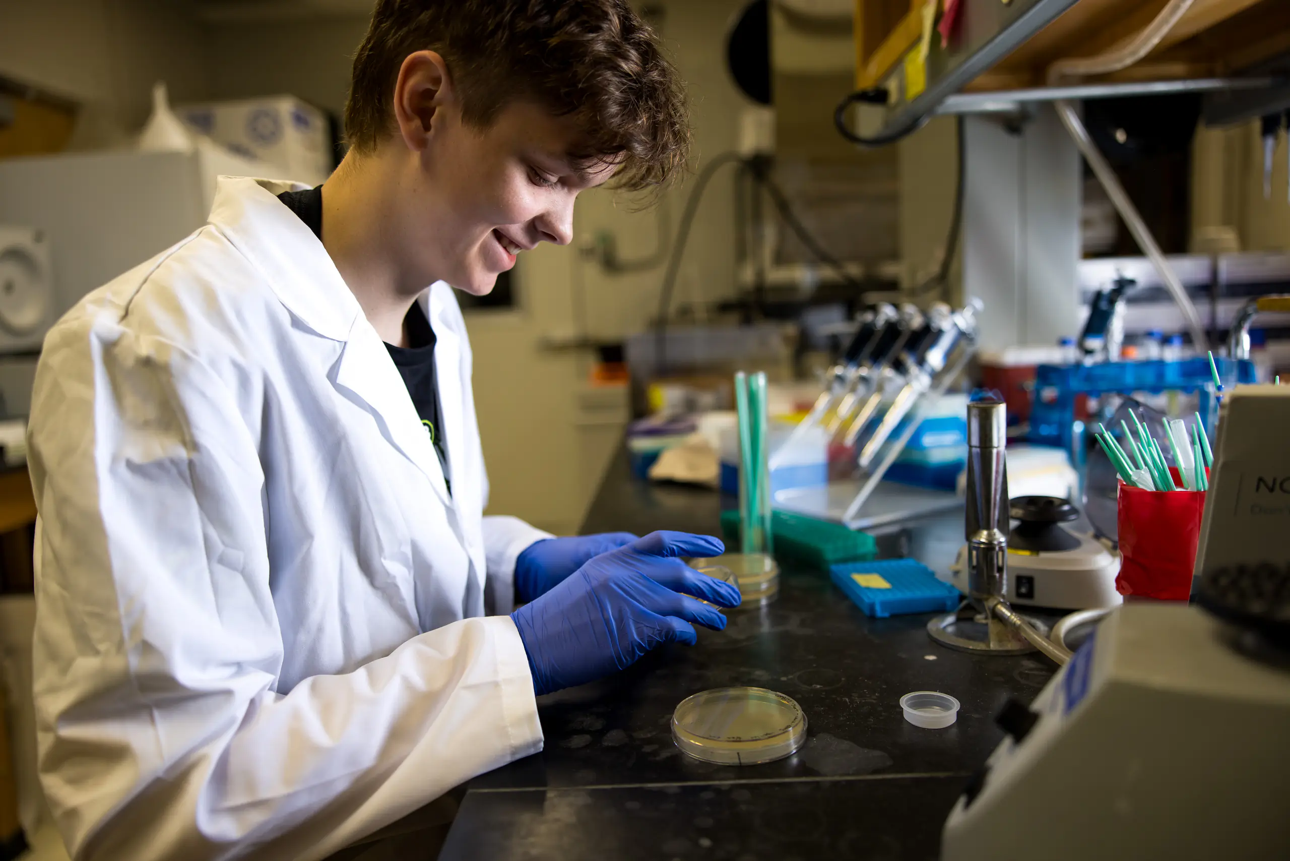 A male student wearing a white lab coat looks at a petri dish in a dim laboratory