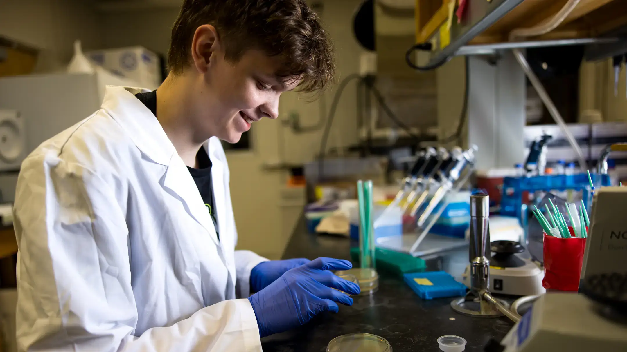 A male student wearing a white lab coat looks at a petri dish in a dim laboratory