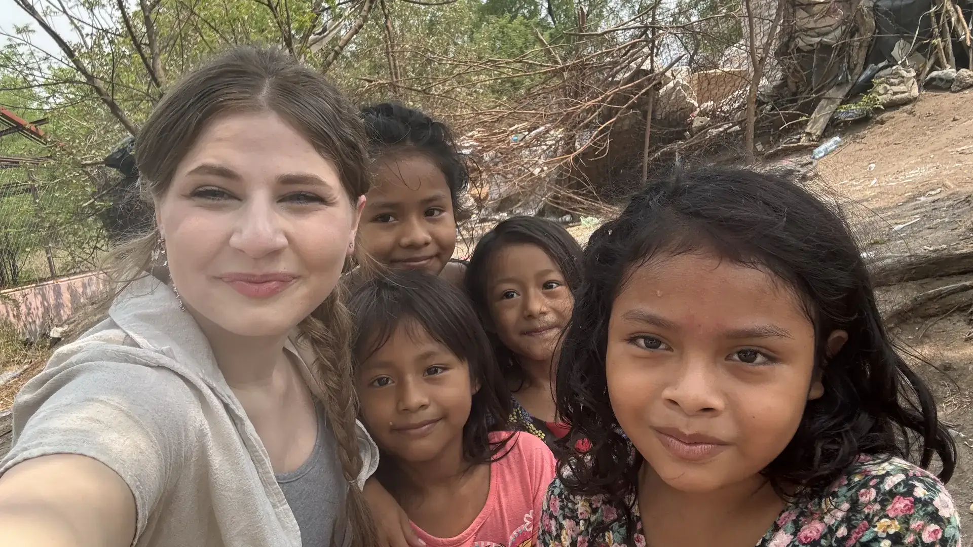 Woman taking a selfie outdoors with four young girls, with trees and makeshift shelters in the background.