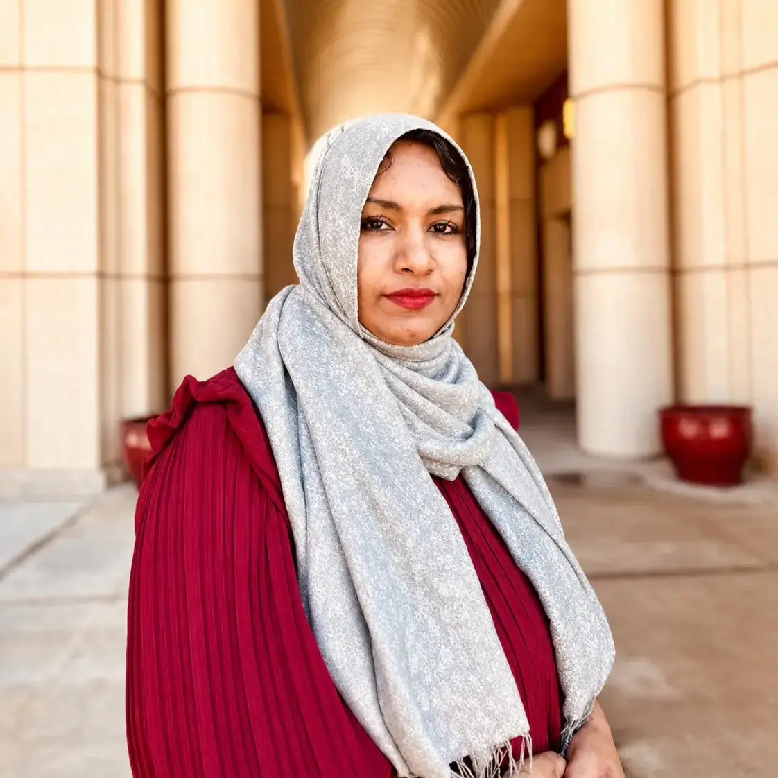 A woman wearing a red dress and a gray headscarf poses in front of a columned architectural structure.