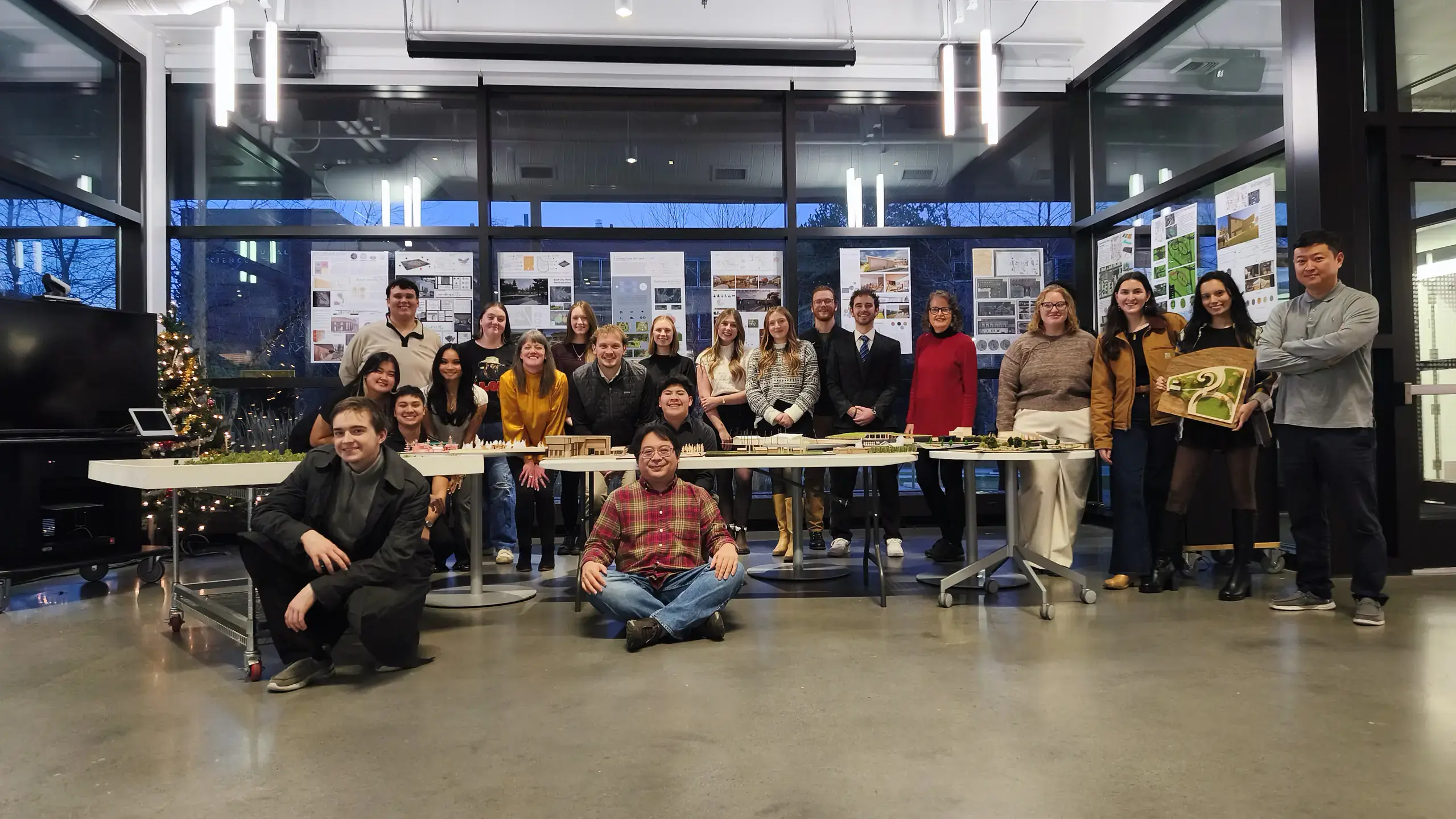 21 people, CAA students and faculty, pose in front of posters and models on tables.