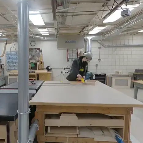 Student with protective gear in woodshop space using tools to work on a wooden frame.