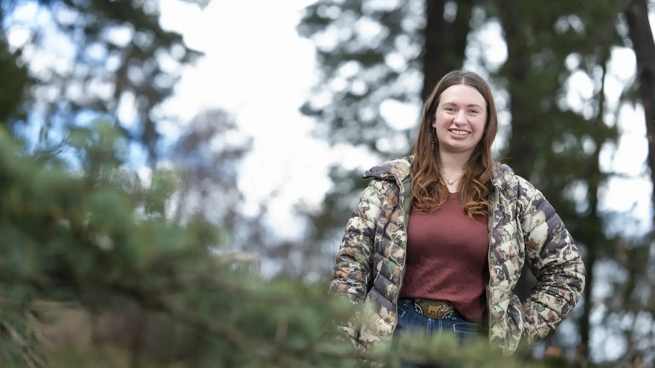 Woman wearing jeans a polo and camouflage jacket stands in a forest. 