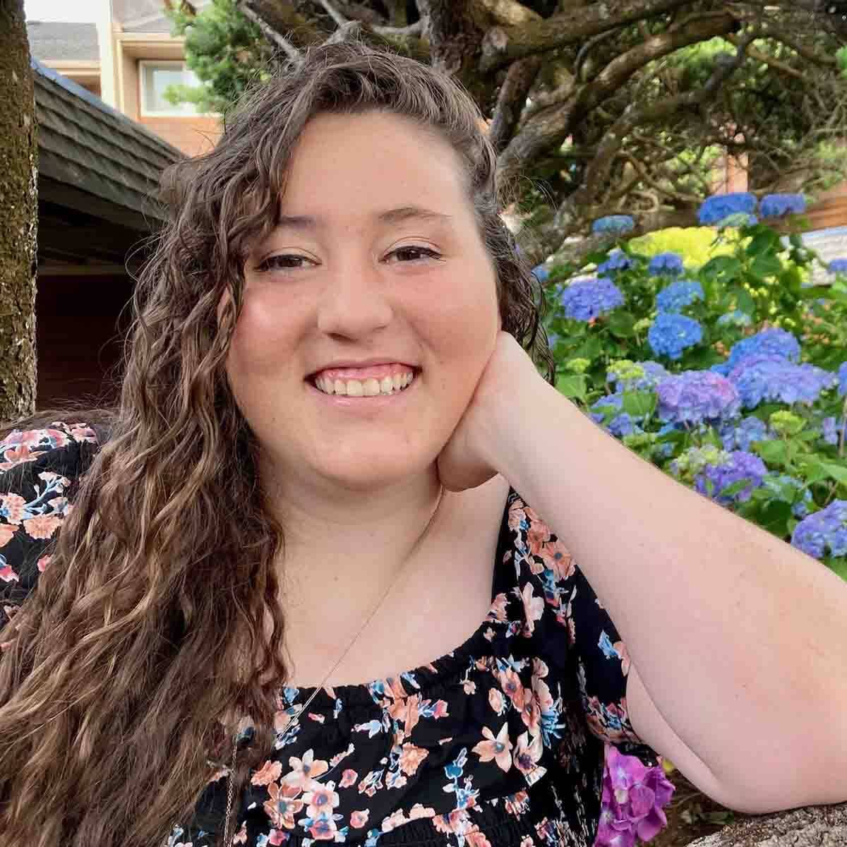 A woman smiling outdoors with hydrangea flowers and trees in the background.