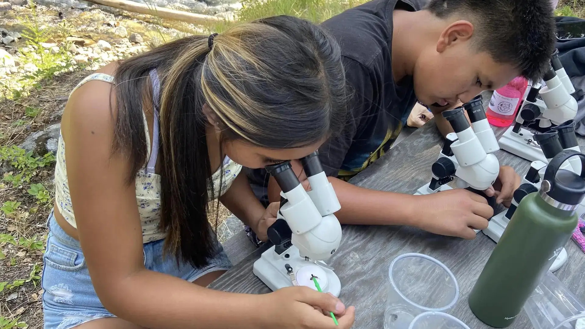 Two youth study specimens through a microscope set up on a picnic table with a cascading stream in the background.