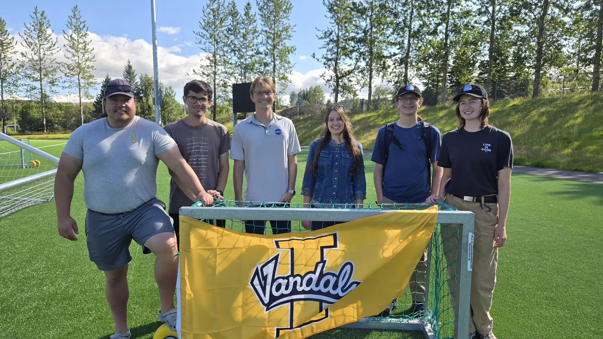 Six students pose on a sports field behind a goal draped with a yellow University of Idaho Vandal banner.