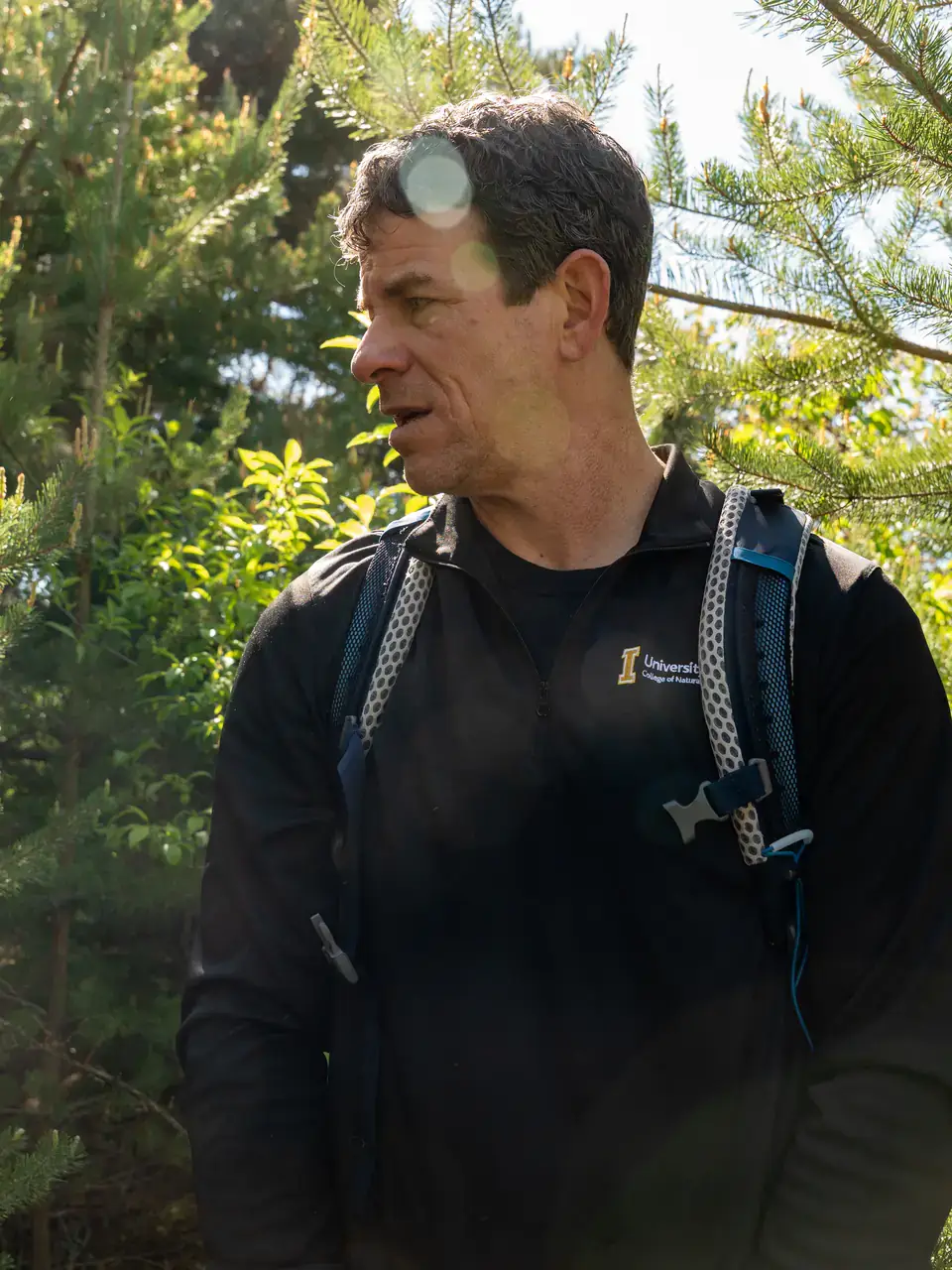 A person wearing a black jacket and a backpack is standing outdoors among green trees, lit by sunlight with some lens flares visible.