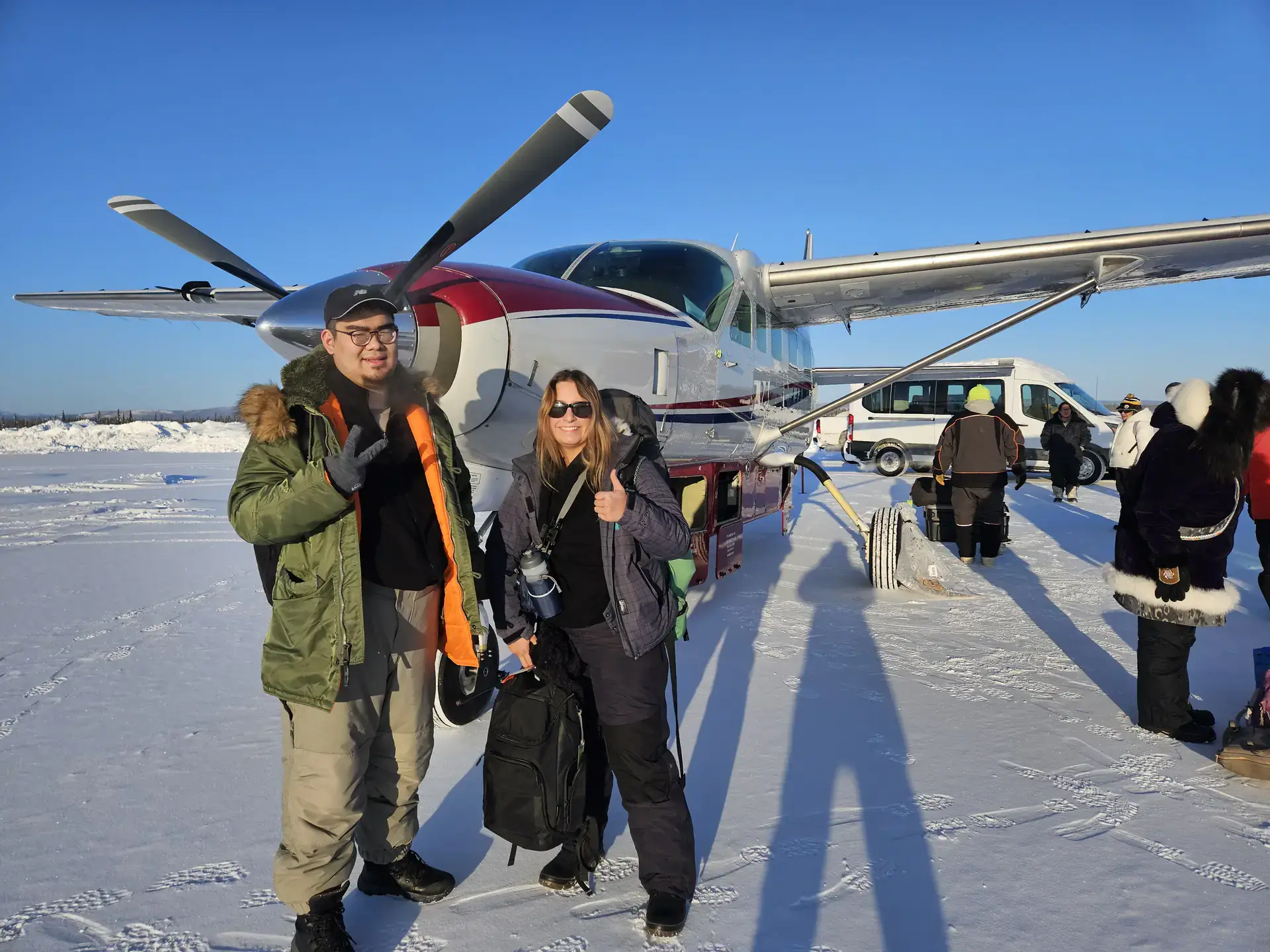Anna Bliss and Young Sim, smiling and wearing winter coats with hoods, in front of a small red and white aircraft, Snow covers the tarmac.