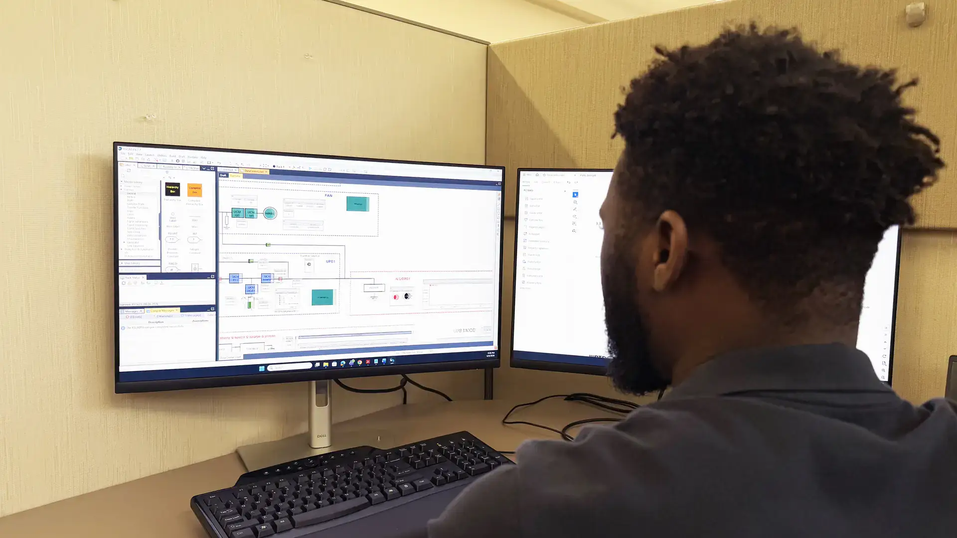 Man working at a cubicle with dual monitors displaying engineering design software.