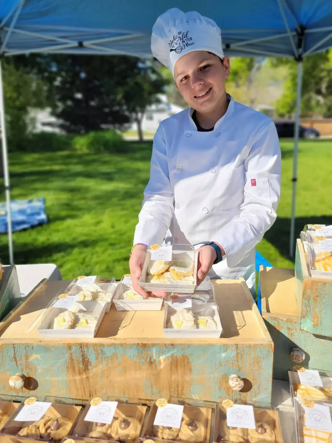 A boy in a chef’s hat and jacket holds a box of icing-covered cookies.