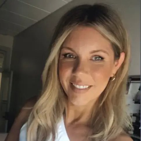 A woman with long blonde hair wearing a white one-shoulder top smiles in a room with neutral colors and soft lighting.