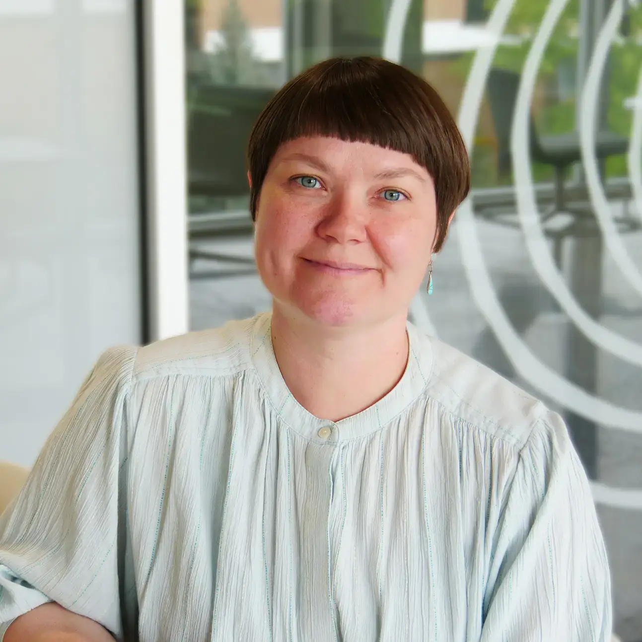A person seated indoors, wearing a light-colored blouse with short hair and earrings, smiling at the camera.