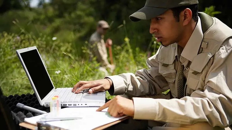 Man outdoors working on laptop taking notes in field