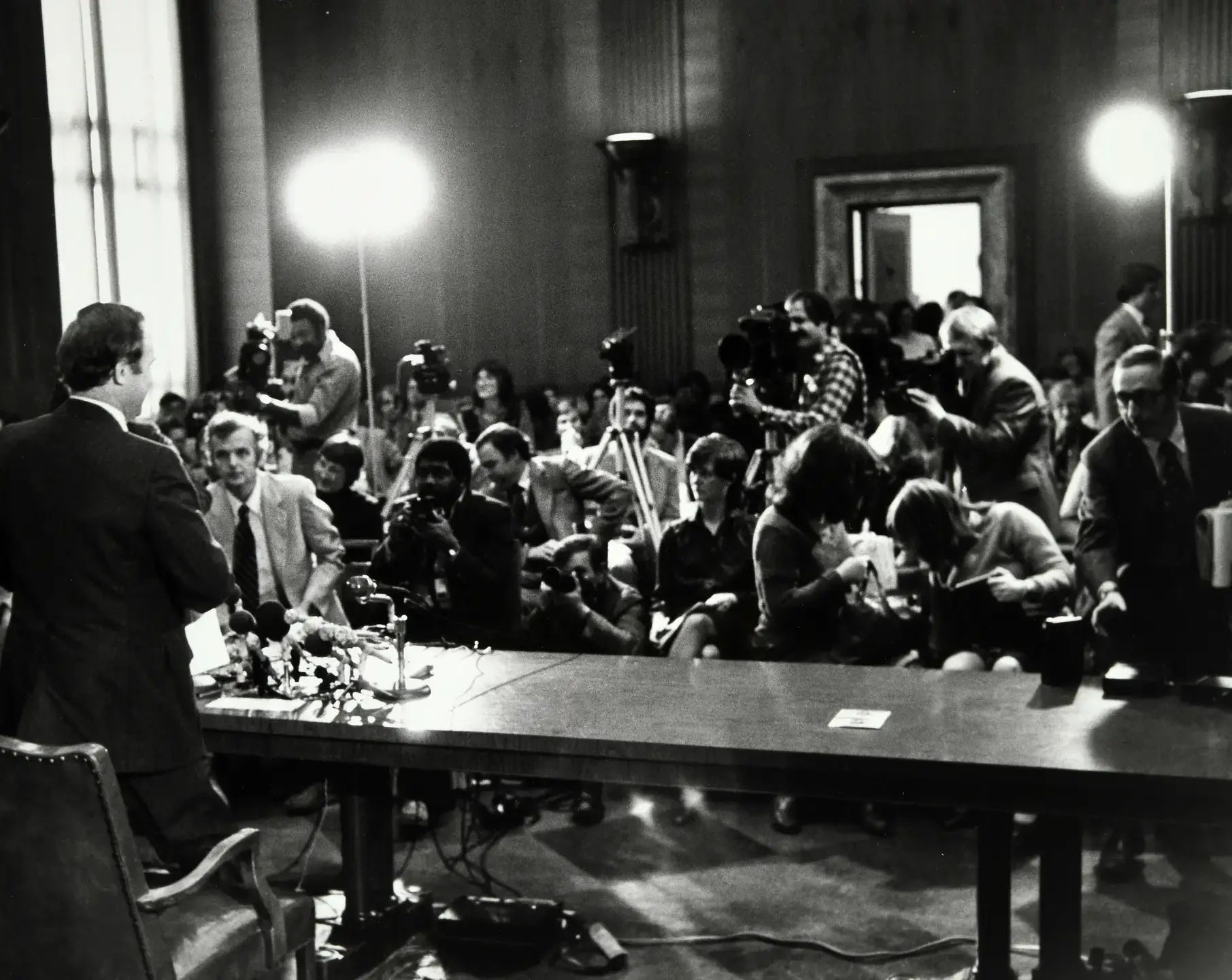 Man speaking at a table with microphones and photographers crowd a hearing room.