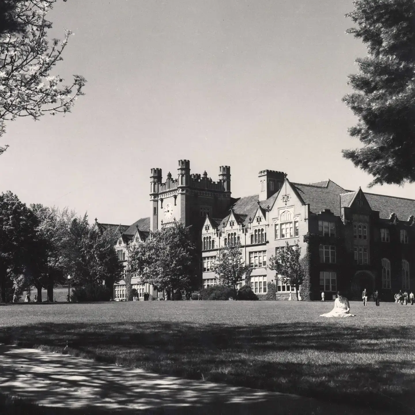Black-and-white photo of a large Gothic-style campus building across a broad lawn, framed by trees.