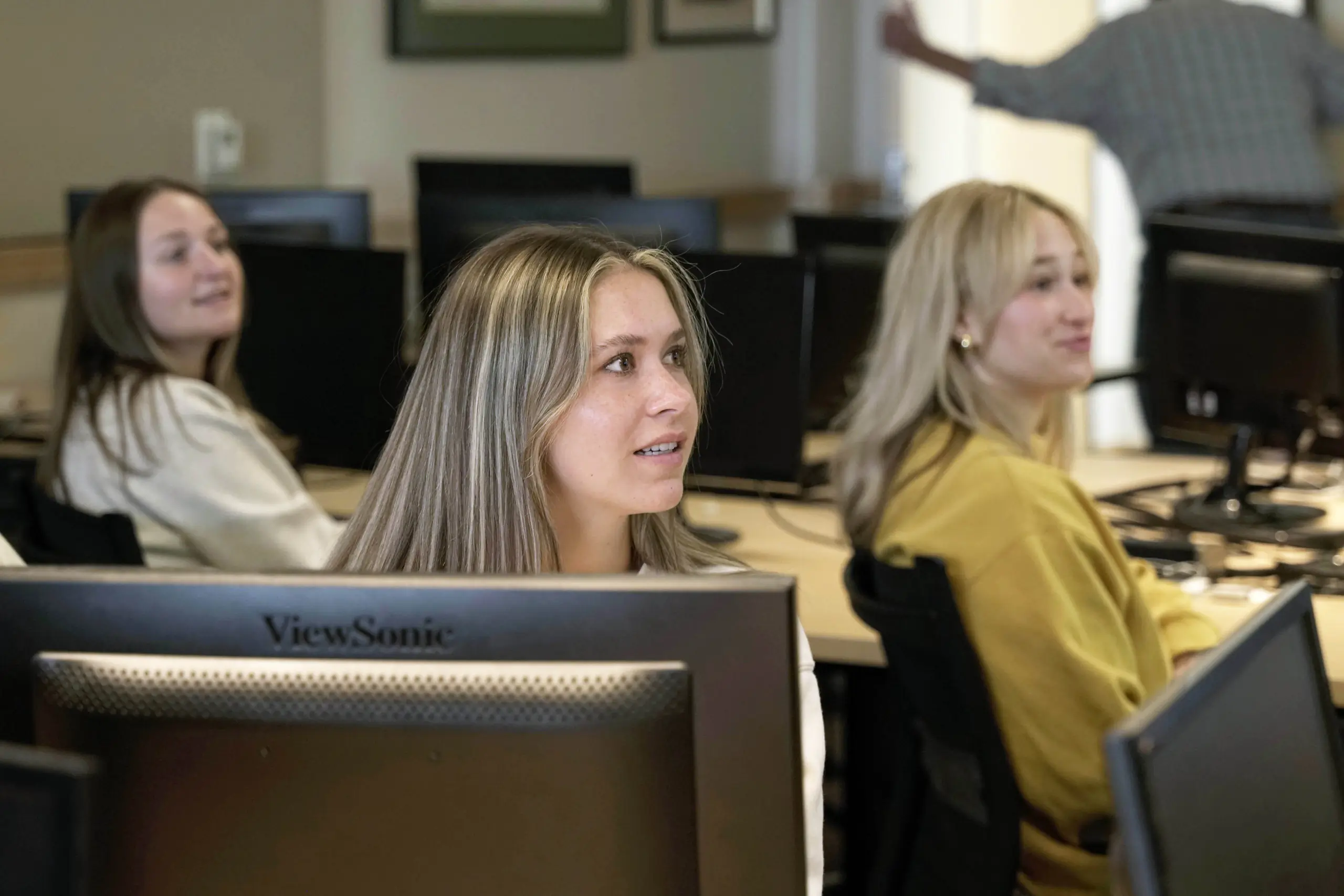 A girl looking to the right in a computer lab with other students.