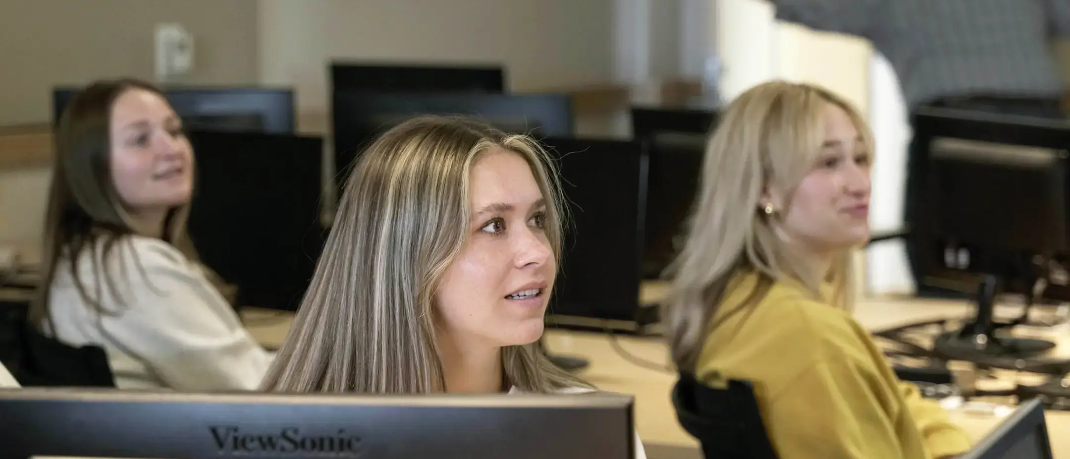 A girl looking to the right in a computer lab with other students.