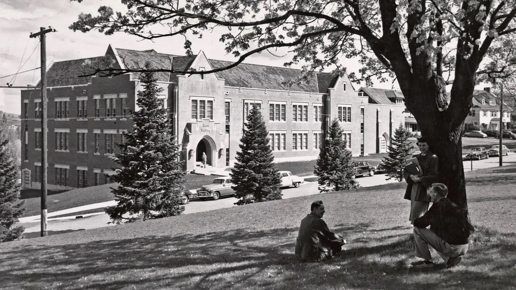 Black-and-white photo of a campus music building, with students sitting under a large tree in the foreground.
