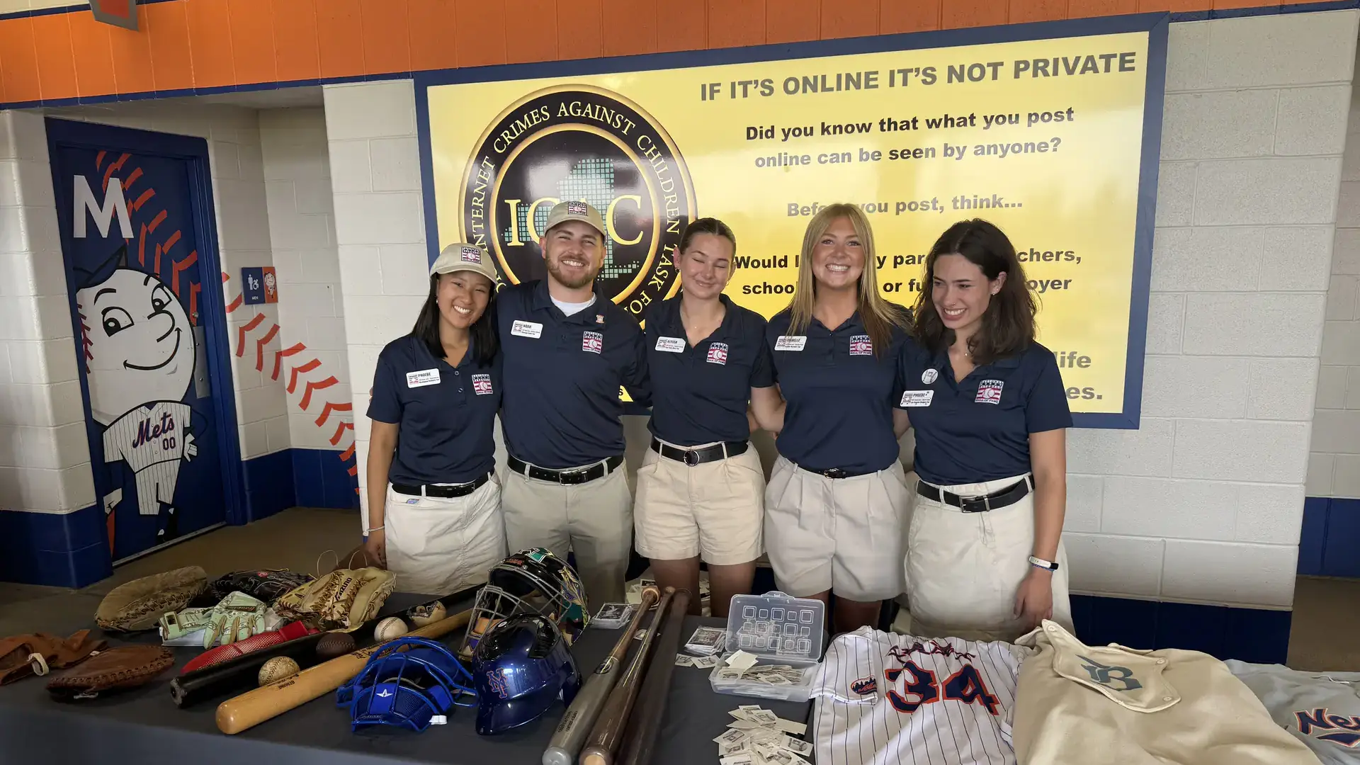 Five people wearing blue shirts and white shorts standing behind two tables.
