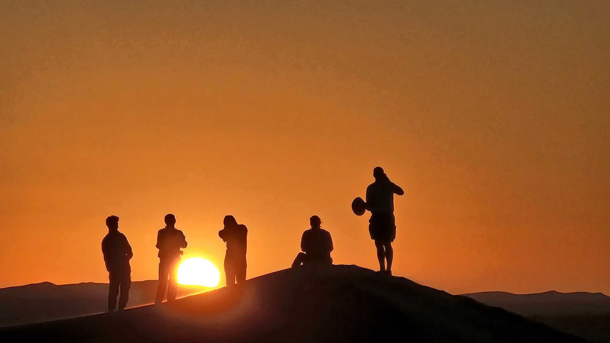 Silhouettes of people atop sand dunes at sunset, with the sun glowing brightly in a clear, orange sky.