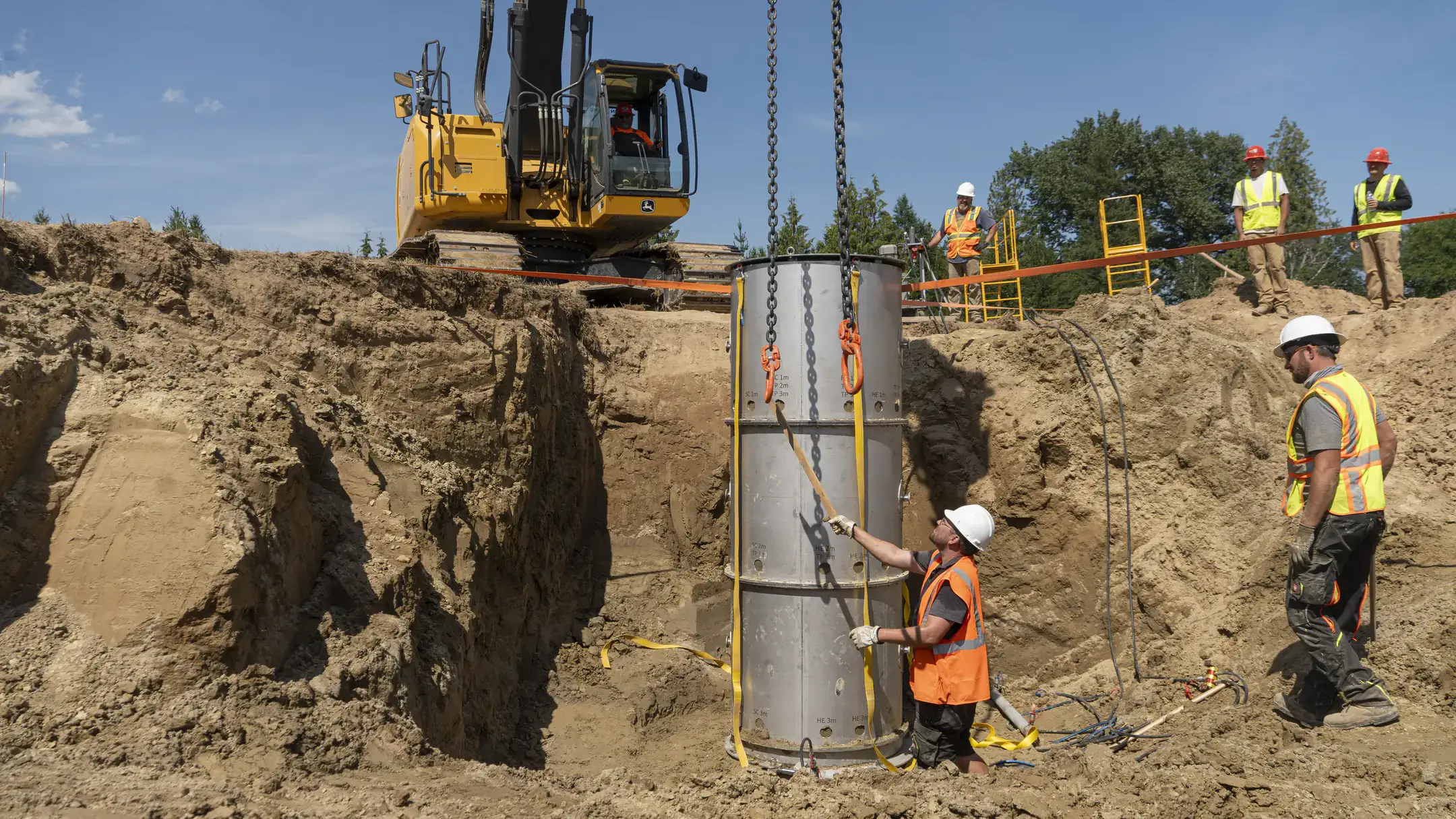 A metal cylinder containing a soil core is lifted using heavy equipment from a dirt wall.