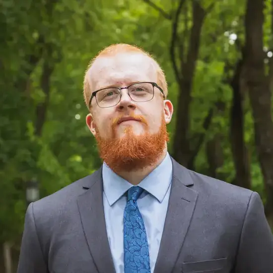 CJ Wagner in a gray suit and blue tie, standing on a tree-lined path outdoors.