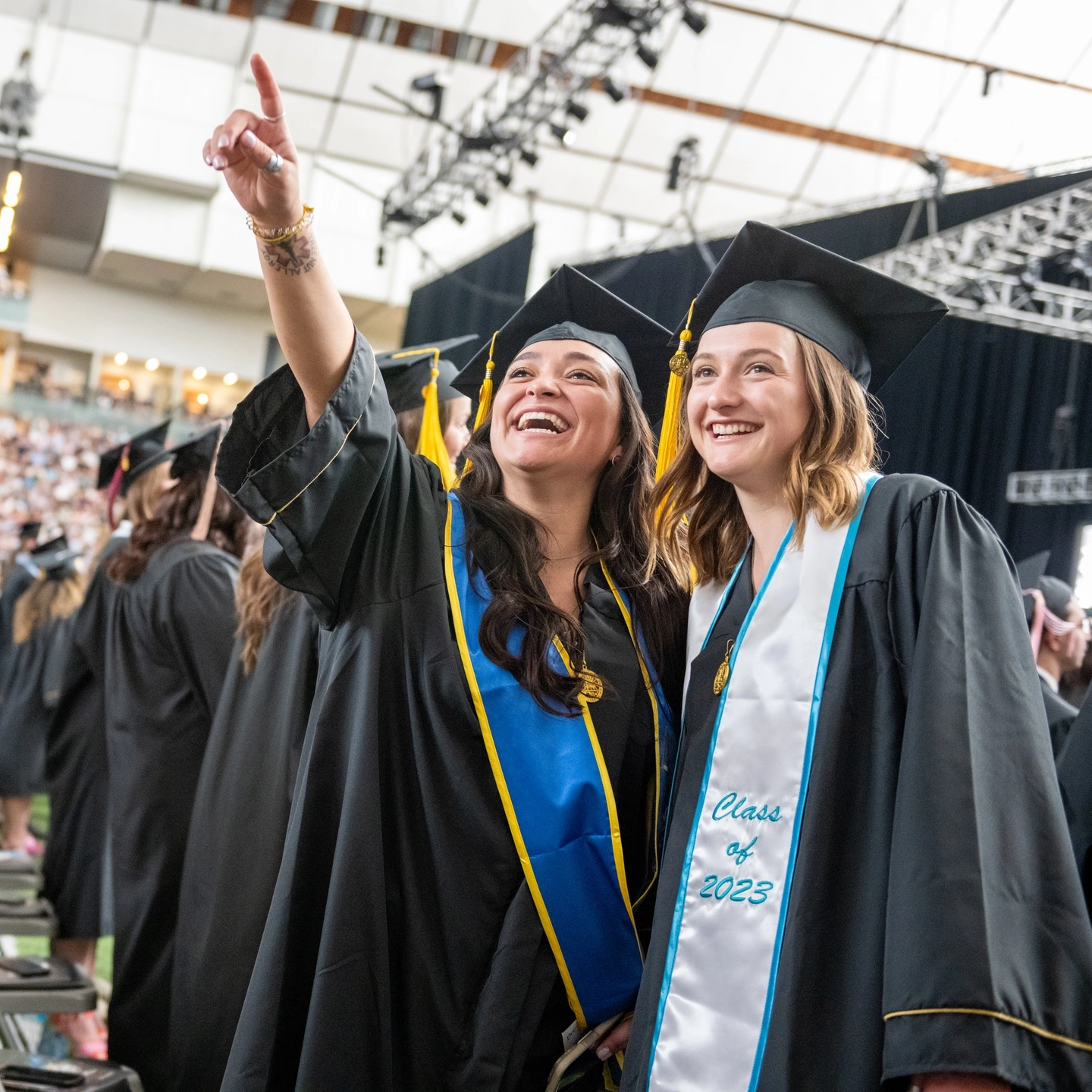 Two graduating students in their cap and gown, with one pointing to the bleachers