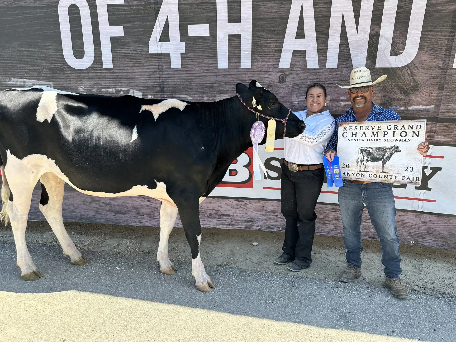 A woman holds a large black-and-white cow by its bridle, standing beside her grandfather, who wears a cowboy hat and holds two blue ribbons and a sign.