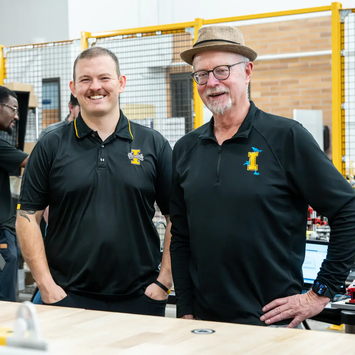 Two men wearing black U of I shirts posing for camera in robotics lab.