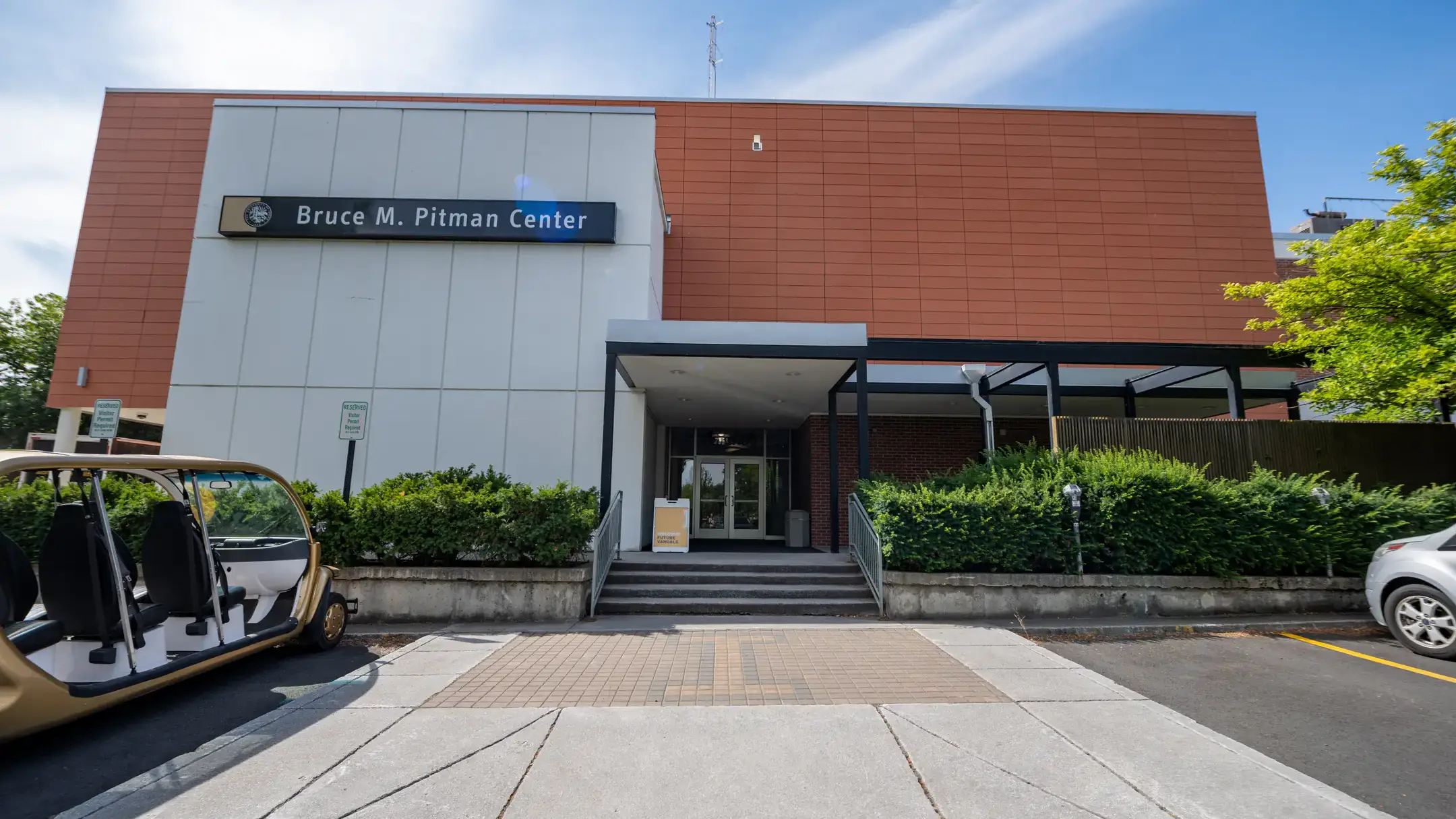 Entrance of the Bruce M. Pitman Center with stairs, a pathway, and nearby greenery on a sunny day.
