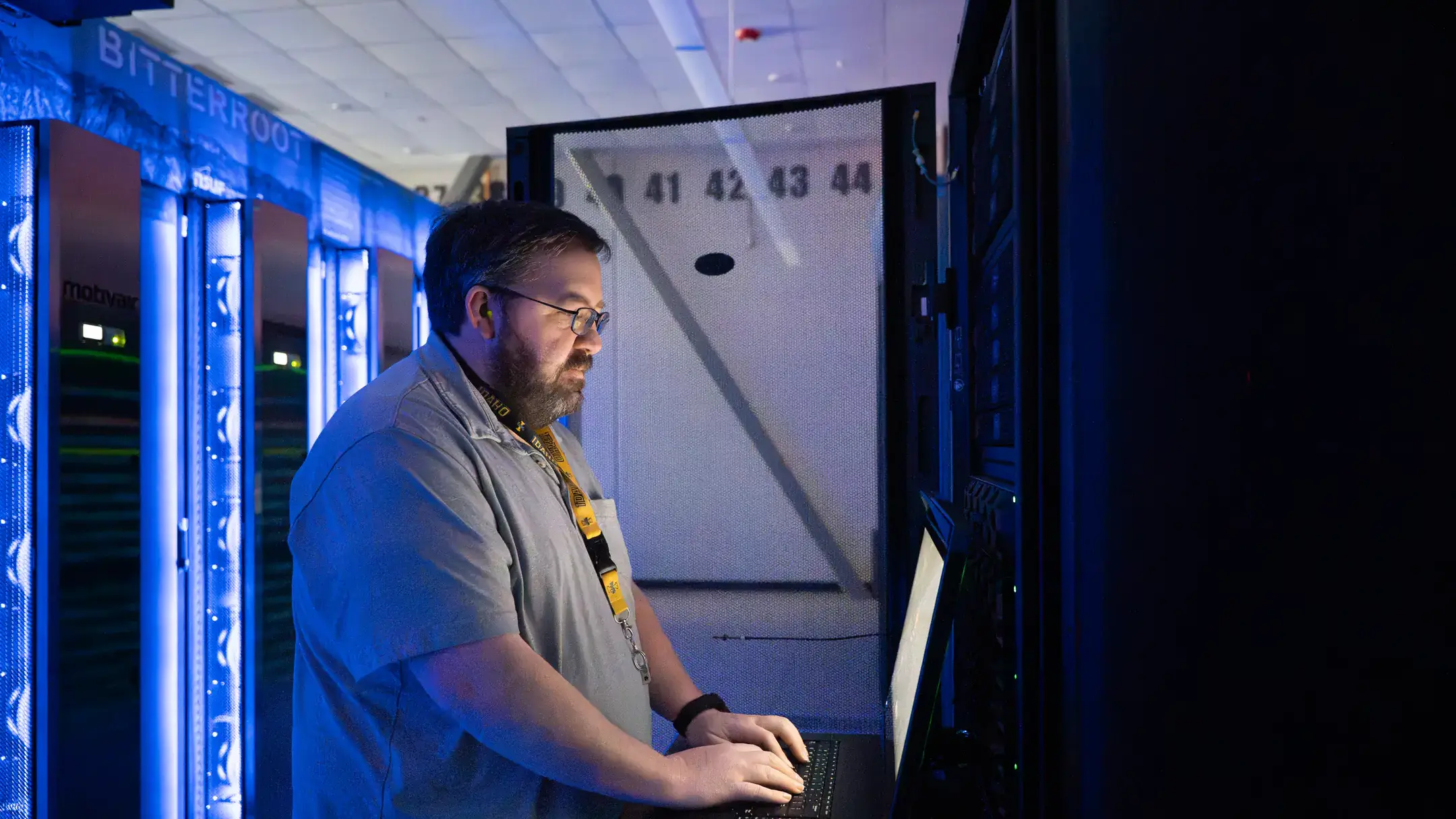 A person working at a computer in a dimly lit room with server equipment illuminated in blue light.