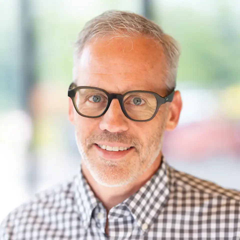 A man with glasses wearing a checkered shirt smiles at the camera with a blurred background.