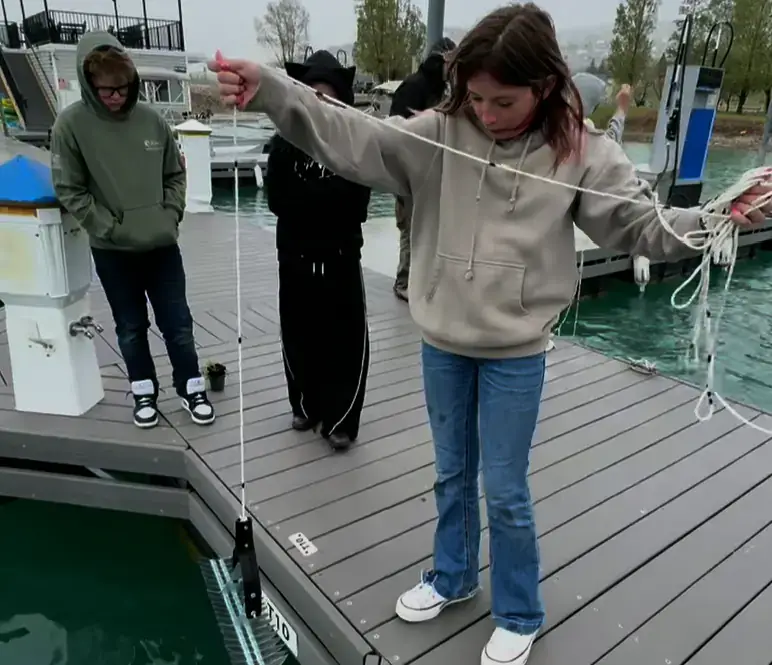 A girl standing on a dock pulls up a rake attached to a long rope from deep within a lake