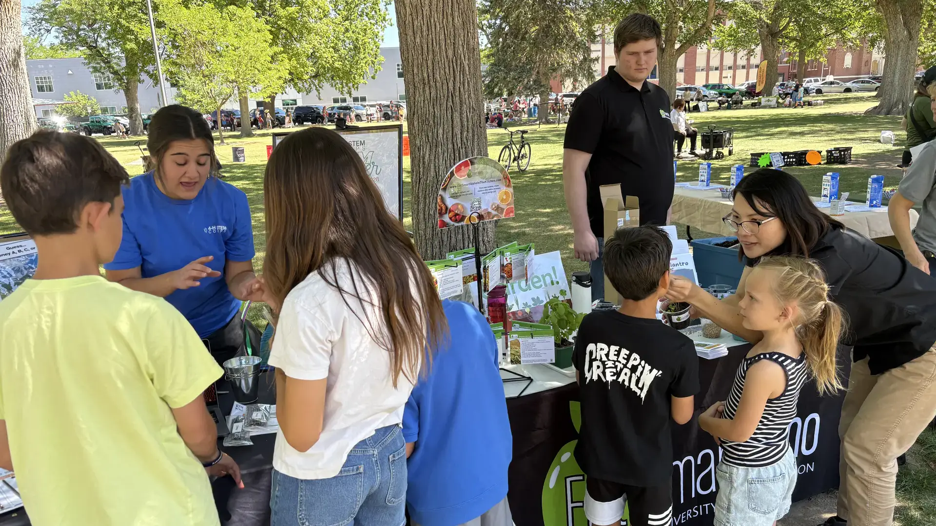 Children receive instructions for educational activities at a booth in a shady park.