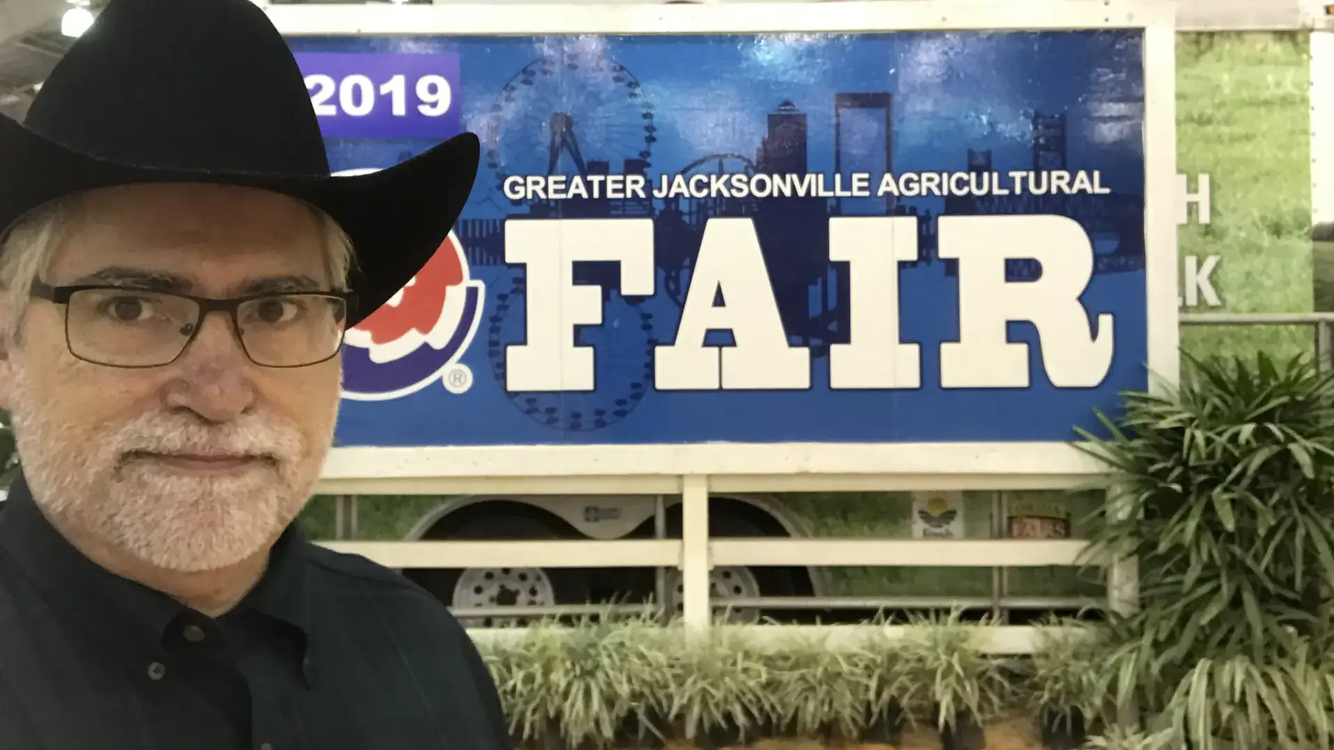 A man wearing a black cowboy hat stands in front of a sign reading 'Greater Jacksonville Agricultural Fair 2019' under an American flag.