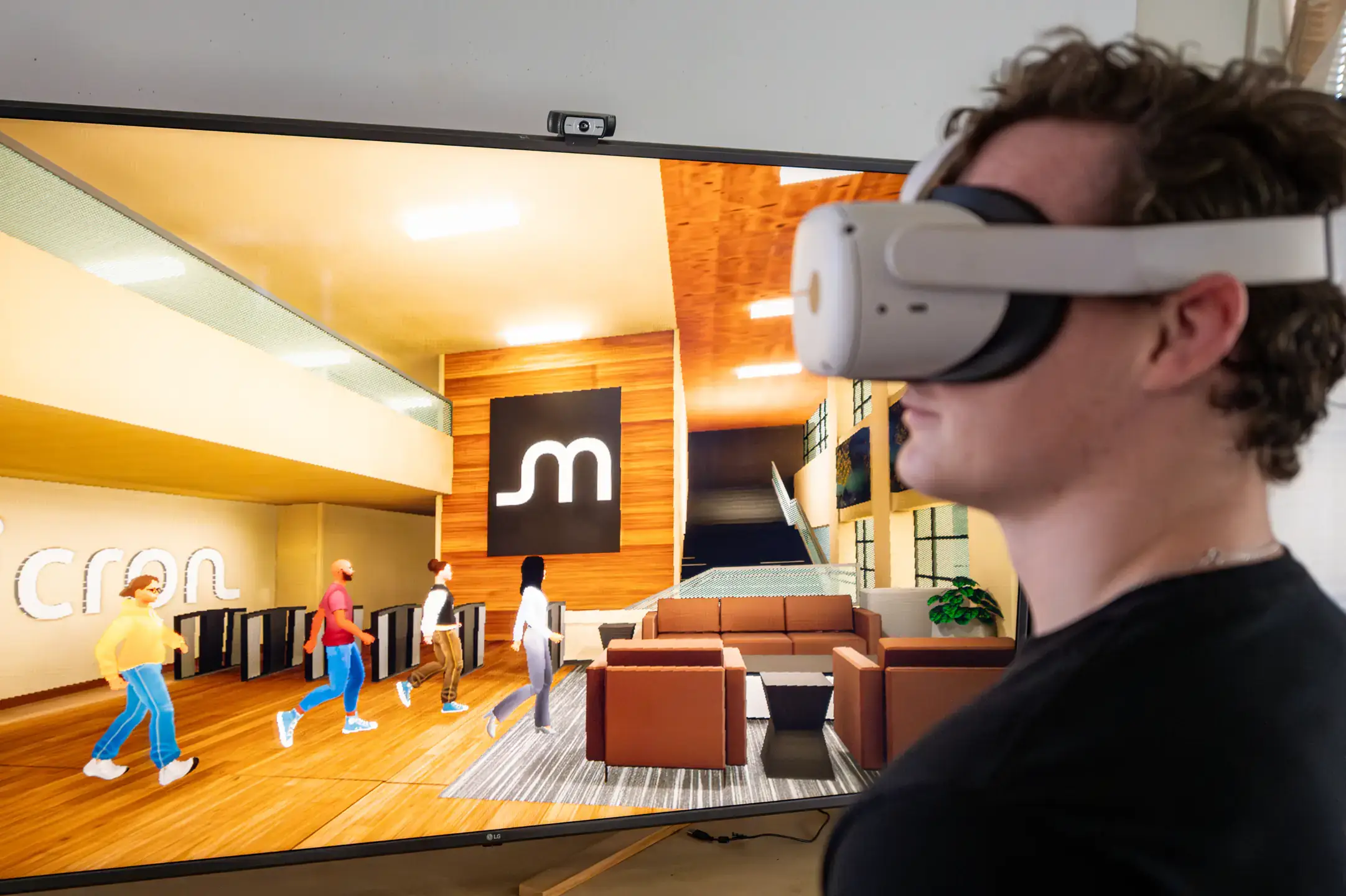 Man wearing a VR headset in front of a tv with four avatars in a virtual Micron room.