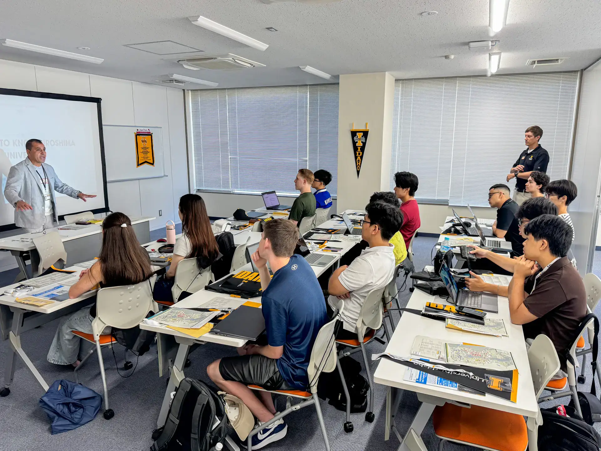 A classroom setting with students participating in a lecture given by a presenter standing at the front near a projector screen.