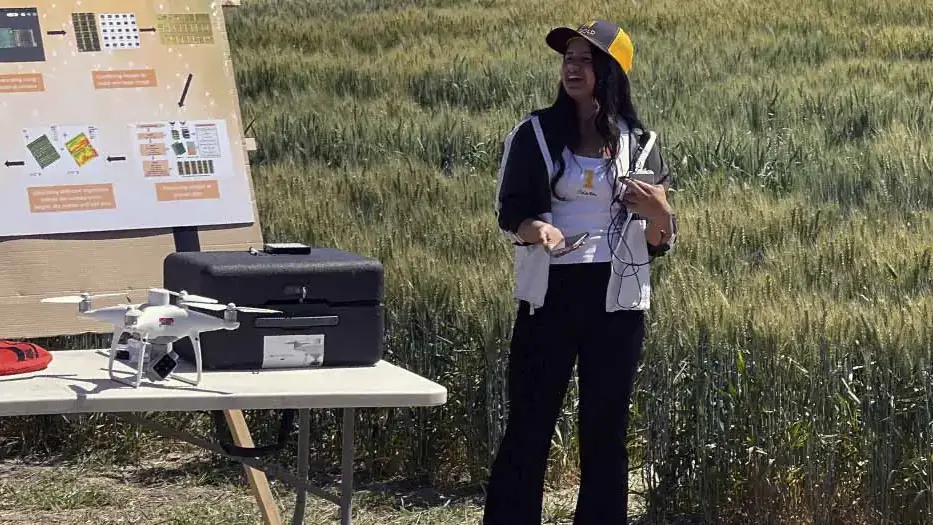Pabitra Joshi stands in a field beside a table with a drone and equipment with a process poster on display in the background.