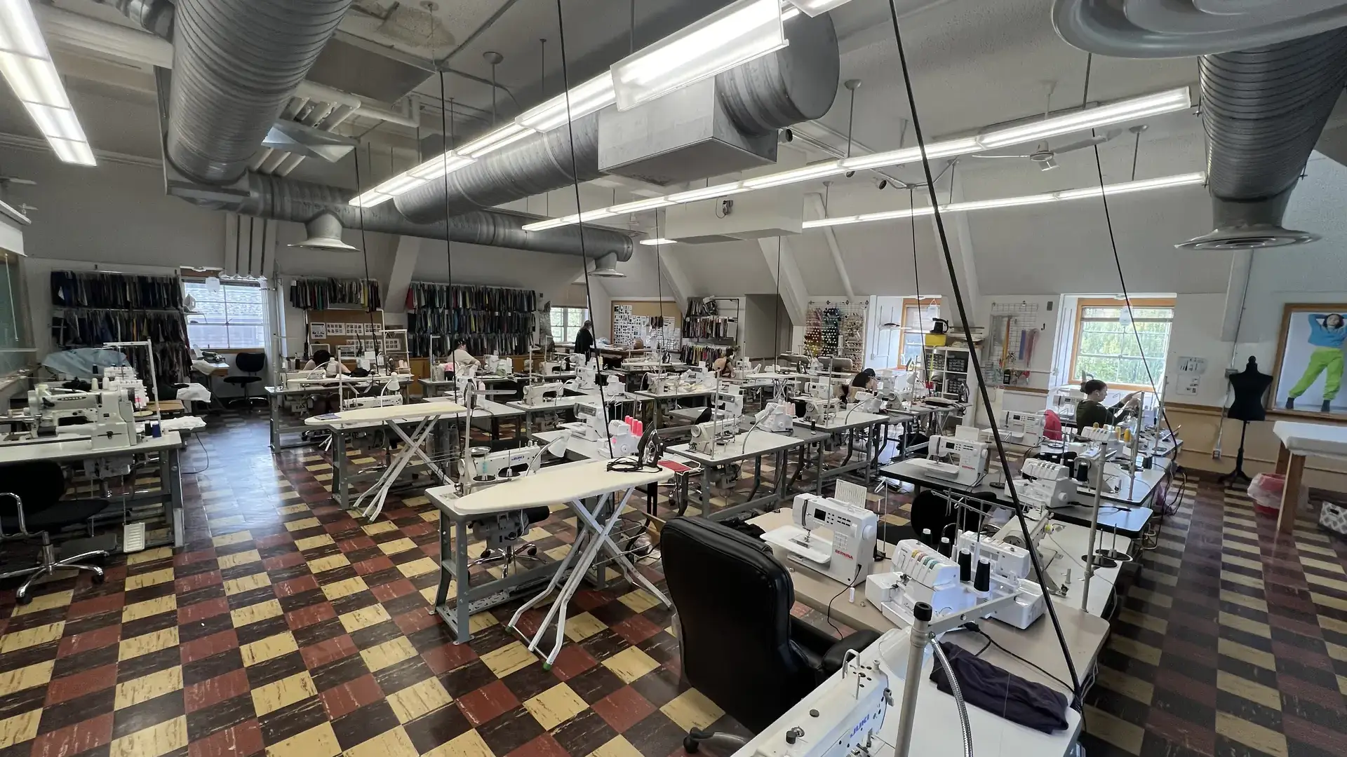 A well-lit textile production studio featuring rows of sewing machines, work tables, and shelves of materials along one wall.
