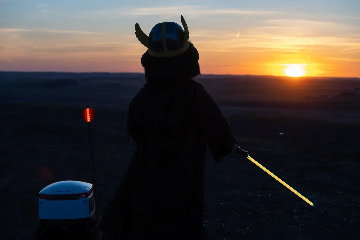 Joe Vandal, in a Jedi robe and holding a yellow lightsaber, stands next to a Starship delivery robot at sunset.