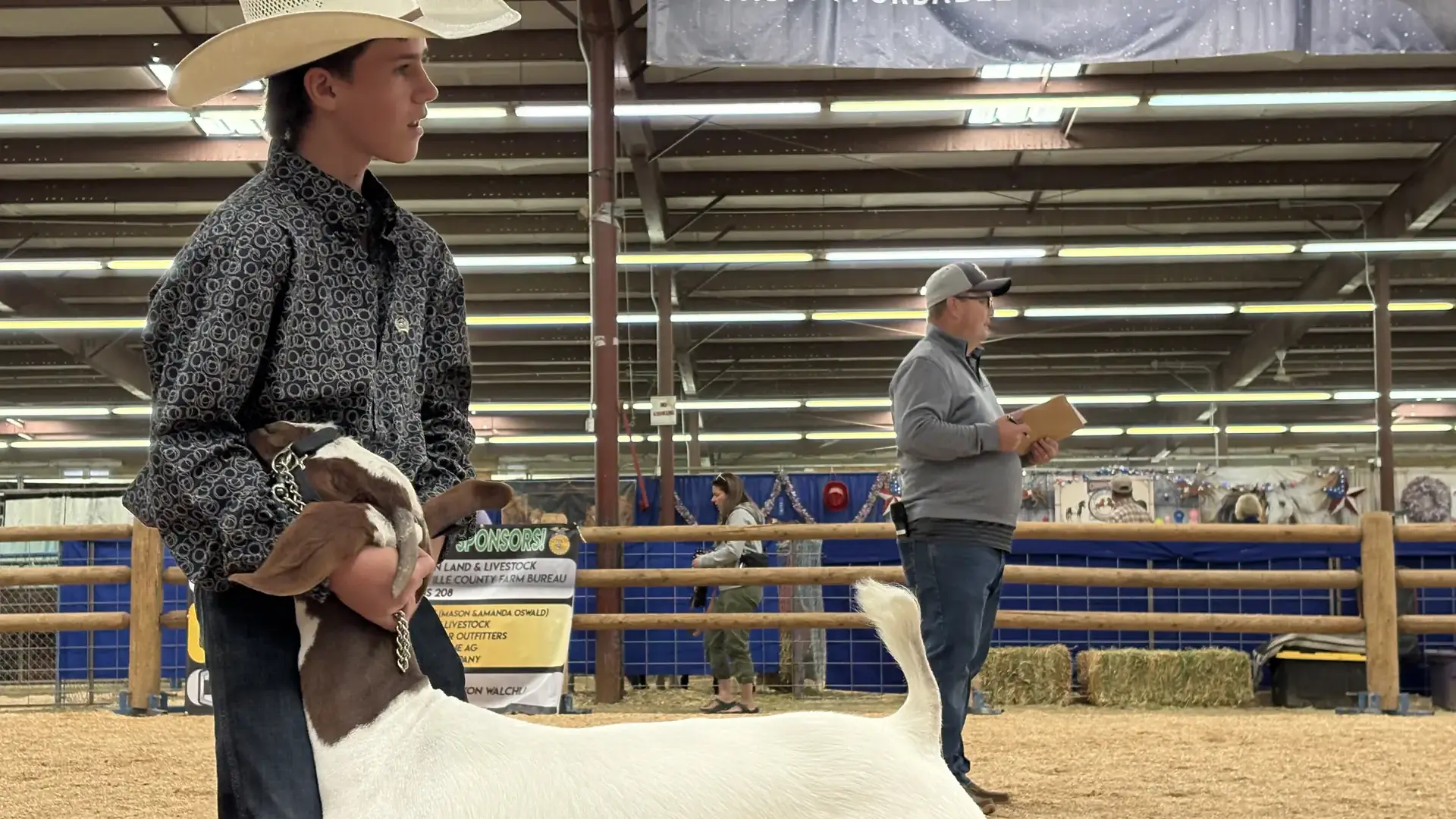 Young person in cowboy hat holding a goat in an indoor fair setting with banners and spectators visible in the background.